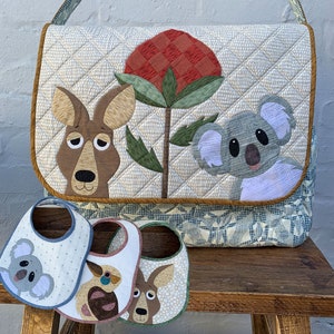 May include: A quilted nappy bag with a kangaroo, koala, and gum blossom design. The bag is beige with a blue and white patterned lining. Three matching bibs are also pictured, each featuring a different animal design.