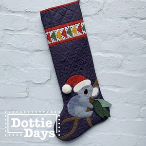 Op de afbeelding: Een paarse, gewatteerde kerstsok met een rood, groen en geel patroonband. De sok heeft een grijze koalabeer die een rode kerstmanmuts draagt en een groen blad vasthoudt.
