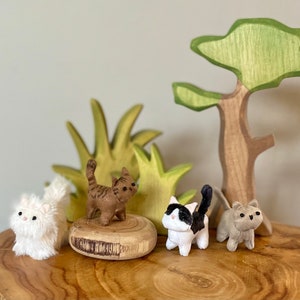 May include: Four plush cat figurines on wooden bases, with a wooden tree and green foliage in the background. The cats are brown and white, black and white, and grey.