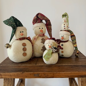 May include: Four white snowman ornaments with different coloured hats and accents. The snowman on the left has a green hat with a bell, the snowman in the middle has a red and white hat, the snowman on the right has a green and white hat, and the snowman in the front has a green and white hat with a floral accent.