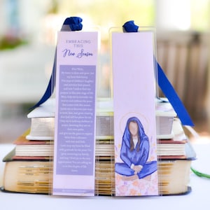 May include: Two bookmarks with blue ribbon ties. One bookmark features the text "EMBRACING THIS New Season" and a prayer. The other bookmark has an illustration of a woman in a blue robe. The bookmarks are resting on a stack of books.
