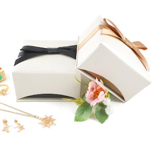 May include: Two white gift boxes with black and brown ribbon bows, a gold necklace with a star pendant, a pair of gold earrings, a gold ring with a green stone, and a gold ring with a white stone. A pink rose with green leaves is in front of the boxes.
