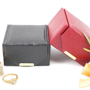 May include: Two small gift boxes, one black and one red, with gold trim.  The red box is decorated with a gold bow and green foliage.  Two gold rings with gemstones are in the foreground.