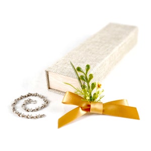 May include: A small, silver chain bracelet with a clasp, a gift box wrapped in beige fabric, and a gold ribbon tied with a bow and green floral embellishments.