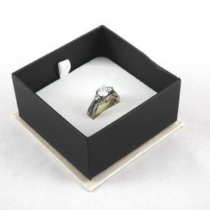 May include: A silver ring with a large clear stone and smaller stones on the band, displayed in a black ring box with a white interior.