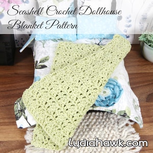 May include: A light green crocheted blanket rests on a dollhouse bed with floral bedding. The blanket is made with a textured stitch pattern. The text "Seashell Crochet Dollhouse Blanket Pattern" is at the top of the image. The website "LydiaHawk.com" is at the bottom.
