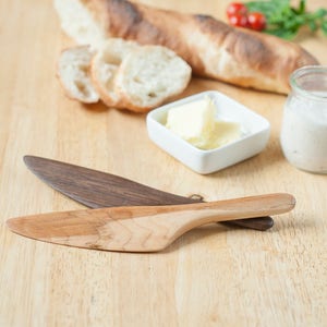 May include: Two wooden butter knives on a wooden surface. One knife is made of dark wood and the other is made of light wood. The knives are in focus and the background is blurred.