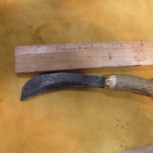 Könnte beinhalten: Ein gebogenes, schwarzes Metallmesser mit einer gemusterten Klinge und einem braunen Hirschhorn-Griff. Das Messer ist etwa 25 cm lang.