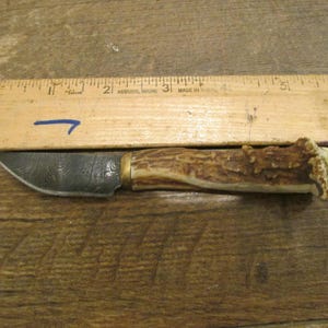 Puede incluir: Un pequeño cuchillo decorativo con una hoja estampada y un mango de asta de ciervo. El cuchillo mide aproximadamente 15 cm de largo, medido con una regla de madera.