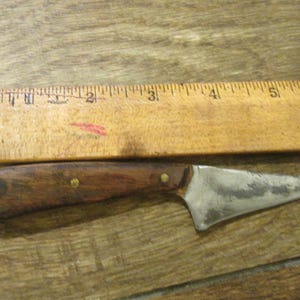 Op de afbeelding: Een klein, handgemaakt mes met een bruin houten handvat en een zilveren lemmet. Het lemmet is ongeveer 10 cm lang en het handvat ongeveer 7,5 cm lang. Het mes ligt op een houten oppervlak.