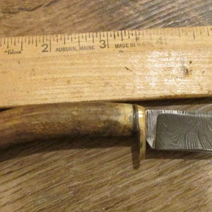 May include: A handmade knife with a patterned steel blade and a handle made from a natural antler. A wooden ruler is placed above the knife. The knife blade is approximately 3 inches long.