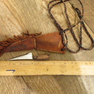 Puede incluir: Un cuchillo pequeño con mango de madera y funda de cuero con flecos. El cuchillo está junto a una regla de madera. La funda es marrón y la hoja del cuchillo es plateada. También hay un cordón trenzado en la imagen.