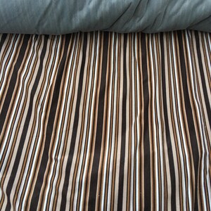 May include: A striped fabric with alternating brown, beige, and white stripes. The fabric is folded in half, with the top half folded over the bottom half.
