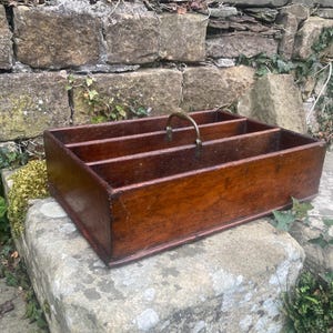 Puede incluir: Un organizador rectangular de madera con cuatro compartimentos y un asa de latón. El organizador está teñido de color marrón oscuro y parece ser vintage. El organizador está sobre una superficie de piedra.