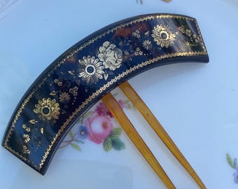 May include: A dark, curved hair comb with gold floral designs. The comb features a black background with gold accents and two amber-colored prongs. The comb is resting on a white plate with floral patterns.