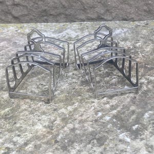 May include: Six silver-coloured metal toast racks. Each rack features a rectangular base, arched supports, and a decorative top. The racks are arranged on a textured, grey surface, suggesting a vintage or antique style.
