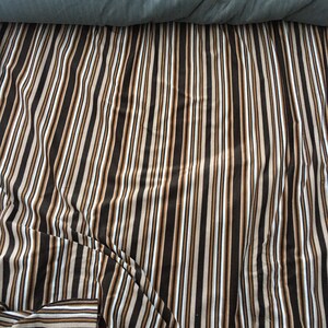 May include: A close-up of a striped fabric with brown, beige, and white stripes. The fabric is folded and creased, creating a textured look.