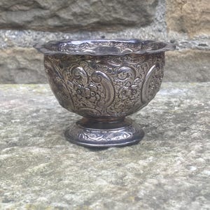 Puede incluir: Un pequeño cuenco plateado ornamentado, con base de pedestal. El cuenco presenta intrincados diseños florales y de cachemira, y un borde festoneado. El cuenco antiguo es probablemente de plata de ley.