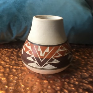 May include: A small, white ceramic vase with a brown and orange geometric pattern. The vase is sitting on a copper-coloured surface.