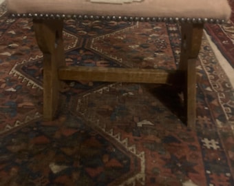 May include: A small wooden stool with a pink upholstered seat featuring a floral needlepoint design. The stool has a simple, X-shaped frame and is sitting on a patterned rug.