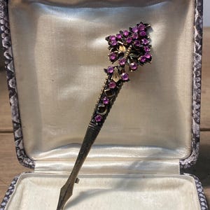 May include: An antique silver-toned hat pin with a decorative floral design. The pin features a cluster of purple gemstones and a detailed stem. It is presented in a vintage, patterned box with a cream-coloured interior.