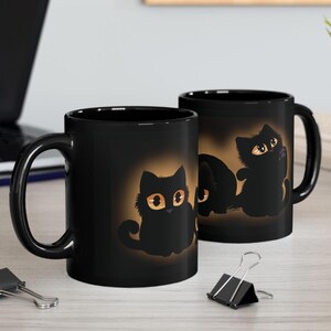 May include: Two black ceramic mugs with a design of three black kittens with orange eyes. The kittens are silhouetted against a light brown background.