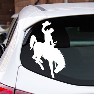 May include: White silhouette decal of a cowboy riding a bucking bronco. The decal is on the back window of a white car.