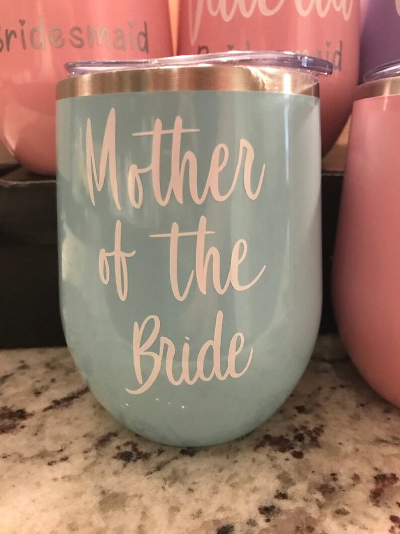 May include: A light blue stainless steel wine tumbler with a gold rim and the text "Mother of the Bride" in white lettering.