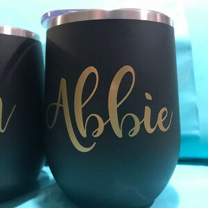 May include: Two black stainless steel tumblers with gold lettering. The tumbler on the left says "Jon" and the tumbler on the right says "Abbie".