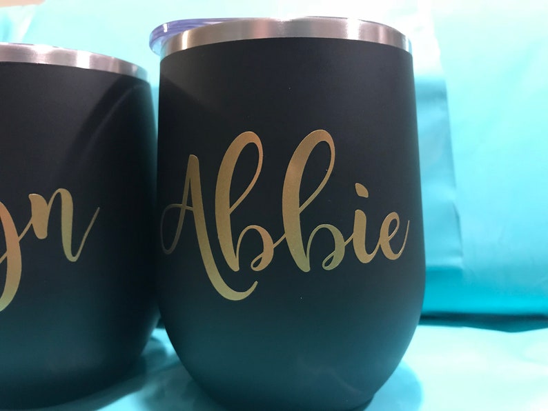 May include: Two black stainless steel tumblers with gold lettering. The tumbler on the left says "Jon" and the tumbler on the right says "Abbie".