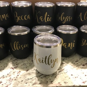 May include: A collection of ten personalized stainless steel wine tumblers with gold lettering. The tumblers are black with the names "Anna", "Becca", "Katie", "Alyssa", "Alison", "Giovanna", "Allison", "Dani", "Stephanie", and "Kaitlyn" written on them. One tumbler is white with the name "Kaitlyn" written on it.