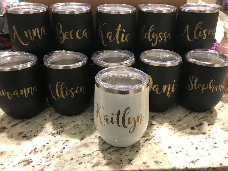 May include: A collection of ten personalized stainless steel wine tumblers with gold lettering. The tumblers are black with the names "Anna", "Becca", "Katie", "Alyssa", "Alison", "Giovanna", "Allison", "Dani", "Stephanie", and "Kaitlyn" written on them. One tumbler is white with the name "Kaitlyn" written on it.