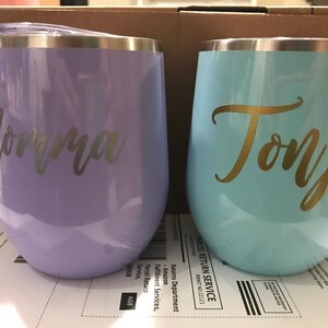 May include: Two stainless steel wine tumblers with personalized names. The tumbler on the left is lavender with the name "Jorma" in silver lettering. The tumbler on the right is light blue with the name "Tonja" in gold lettering.