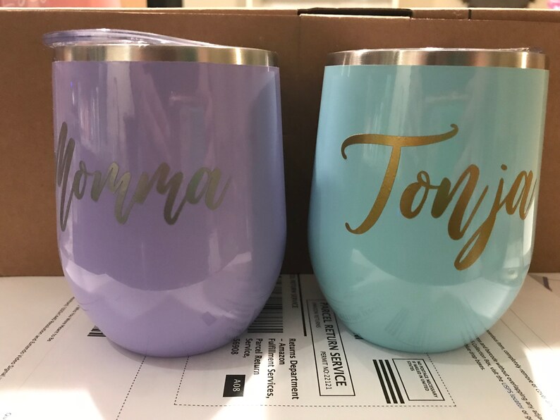 May include: Two stainless steel wine tumblers with personalized names. The tumbler on the left is lavender with the name "Jorma" in silver lettering. The tumbler on the right is light blue with the name "Tonja" in gold lettering.
