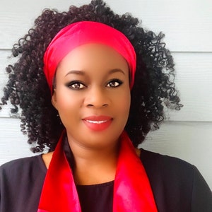 May include: A woman with curly black hair is wearing a black top and a bright pink satin headband and scarf. The scarf is tied around her neck and hangs down her chest.