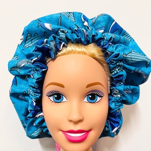 May include: A blue satin bonnet with a white and blue shark print. The bonnet is worn on a doll's head.