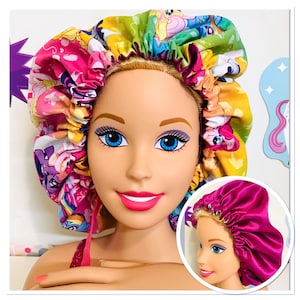 Op de afbeelding: Een roze en paarse satijnen haarbonnet met een kleurrijke My Little Pony print. De bonnet wordt gedragen door een pop met blond haar en blauwe ogen.
