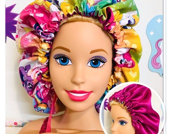 Op de afbeelding: Een roze en paarse satijnen haarbonnet met een kleurrijke My Little Pony print. De bonnet wordt gedragen door een pop met blond haar en blauwe ogen.