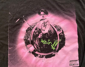 May include: A black t-shirt with a pink and black graphic print of a person in a distorted fisheye lens view. The text "We in" is written in green on the graphic. The t-shirt has a parental advisory label with the text "Explicit Content".