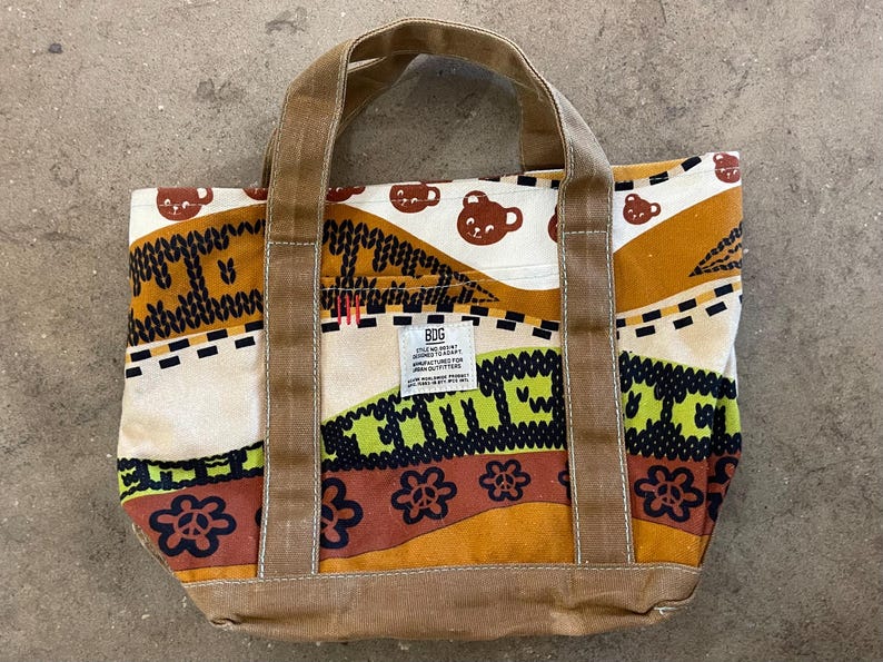 May include: A colorful tote bag with brown handles and a patterned design. The bag features a repeating pattern of brown bear faces, flowers, and geometric shapes in shades of brown, green, and cream. The bag has a label that says "BDG".