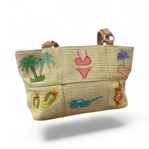May include: Beige straw tote bag with beach-themed embroidered patches. Features palm trees, a pink polka-dot bikini, sunglasses, flip-flops, a cocktail, and an umbrella. Includes tan leather handles and silver metal rings.