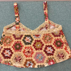 May include: A handmade, floral print top with a yellow background and a honeycomb pattern. The top features a V-neck design, delicate lace trim, and matching straps. The fabric showcases red, green, and white floral details.