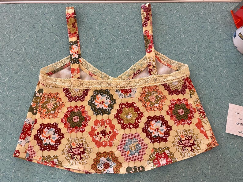 May include: A handmade, floral print top with a yellow background and a honeycomb pattern. The top features a V-neck design, delicate lace trim, and matching straps. The fabric showcases red, green, and white floral details.