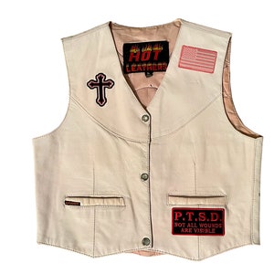 May include: A cream-colored leather vest with a cross, American flag, and "P.T.S.D. Not all wounds are visible" patches. The vest has a zipper closure and two pockets.