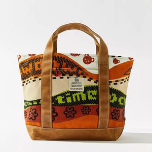 May include: A canvas tote bag with a colorful pattern. The bag features a brown base and handles, with a cream-colored body adorned with orange, green, and red designs. The text "COME" is visible on the front.