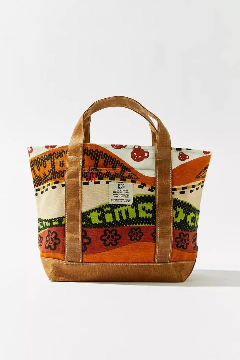 May include: A canvas tote bag with a colorful pattern. The bag features a brown base and handles, with a cream-colored body adorned with orange, green, and red designs. The text "COME" is visible on the front.