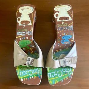 May include: Pair of open-toe sandals with a tan leather strap and buckle. The footbed is brown with colorful illustrations and the words "Rio Coco Beans". The heel features a coffee cup design.
