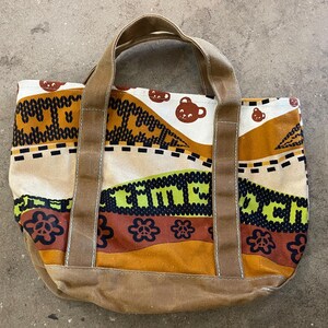 May include: A canvas tote bag with a colorful pattern and brown accents. The bag features a repeating pattern of bear faces, floral designs, and the word "MACH" in green. The handles and base are a solid brown.