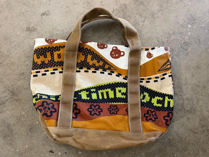 May include: A canvas tote bag with a colorful pattern and brown accents. The bag features a repeating pattern of bear faces, floral designs, and the word "MACH" in green. The handles and base are a solid brown.