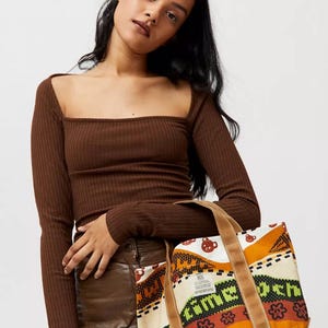 May include: Brown tote bag with a colorful patterned design, featuring orange, green, and white elements. The bag has brown handles and a brown base. The model is wearing a brown long-sleeve top.